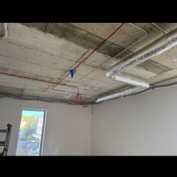 SecVolt Adrian Krupecki - Betonowy sufit z widocznymi przewodami elektrycznymi w kolorze czerwonym i białym, rurami wentylacyjnymi w srebrnej izolacji oraz puszką instalacyjną w kolorze niebieskim.