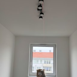 SecVolt Adrian Krupecki - Zamontowane czarne reflektory sufitowe z czerwoną linią lasera w nowym, jasnym pokoju. Widoczna drabina i okno z widokiem na budynek.