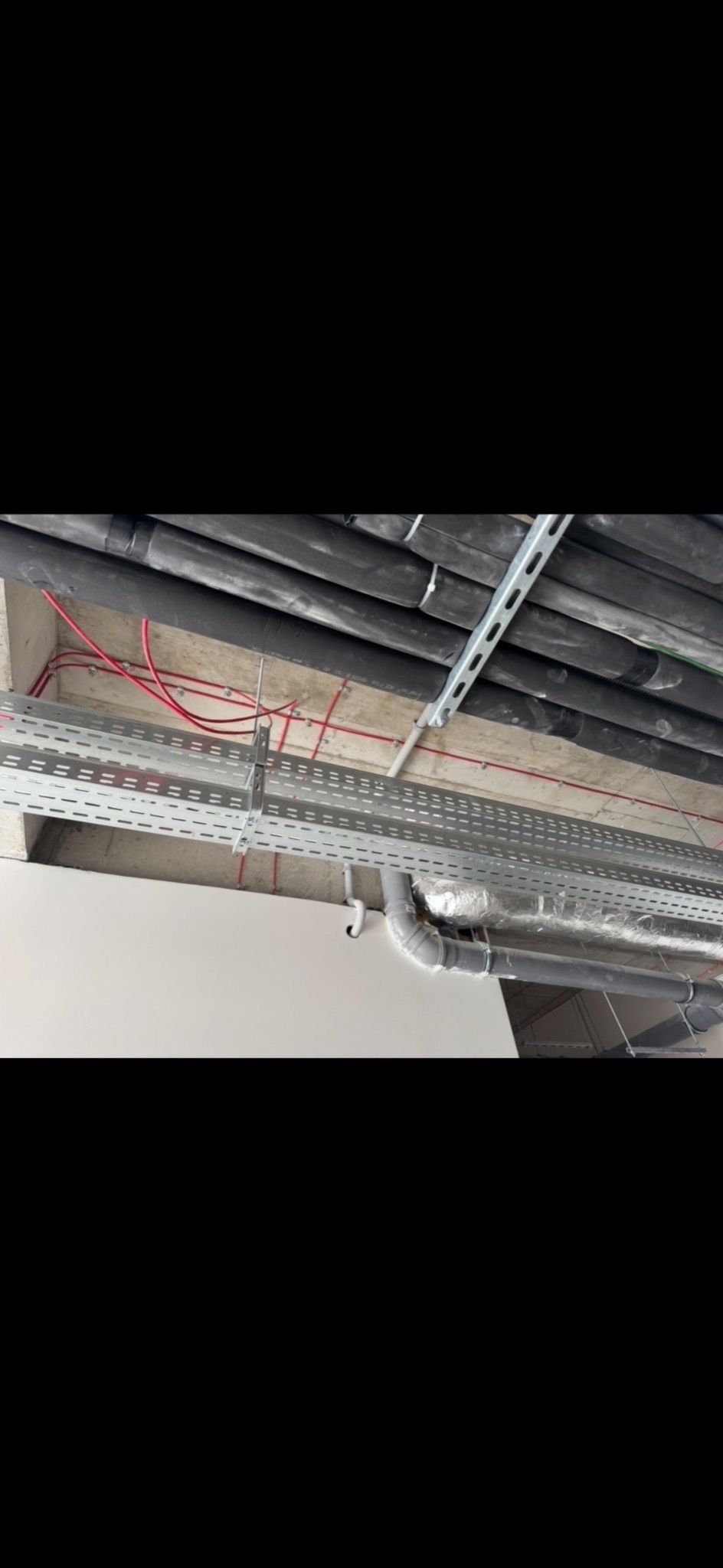 Sufit z widocznymi instalacjami: rury w izolacji, koryta kablowe z przewodami. Szare elementy mocowane do sufitu. Widok z dołu, perspektywa zbieżna.