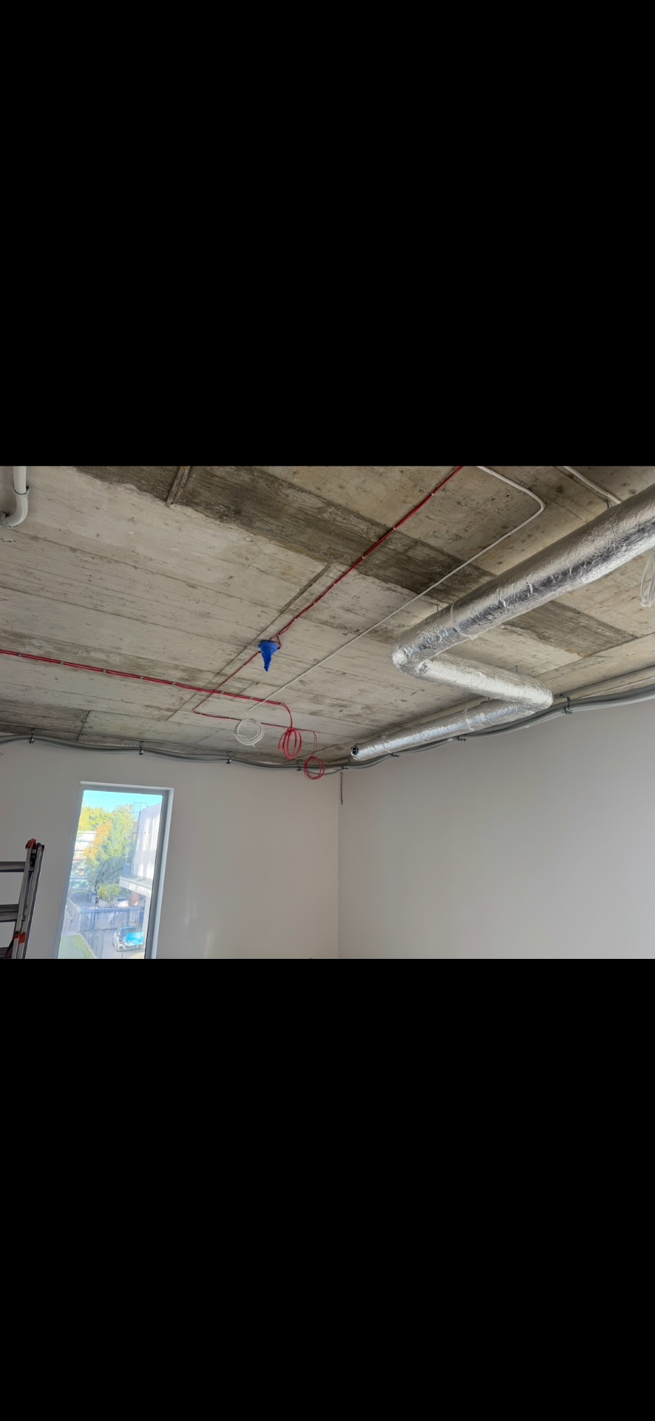 Betonowy sufit z widocznymi przewodami elektrycznymi w kolorze czerwonym i białym, rurami wentylacyjnymi w srebrnej izolacji oraz puszką instalacyjną w kolorze niebieskim.