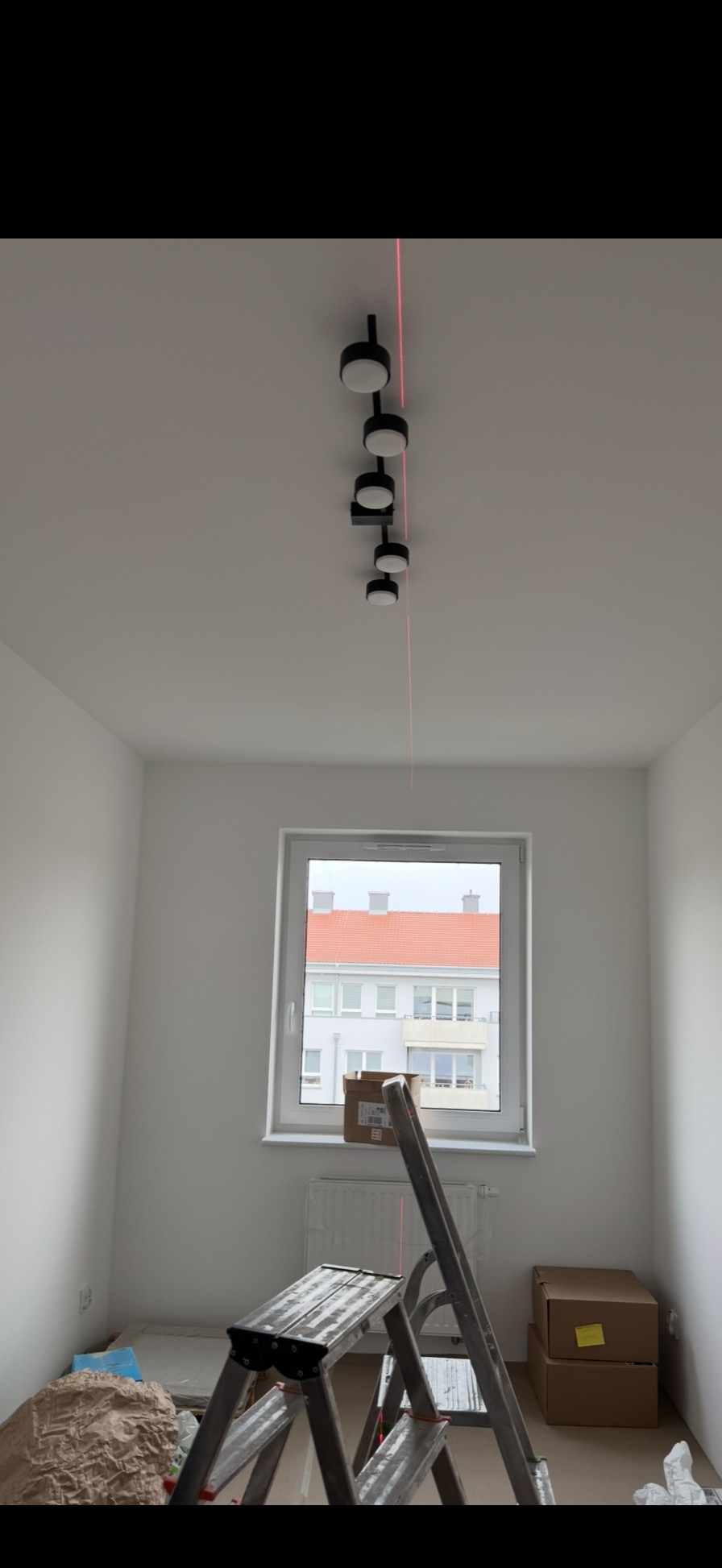 Zamontowane czarne reflektory sufitowe z czerwoną linią lasera w nowym, jasnym pokoju. Widoczna drabina i okno z widokiem na budynek.