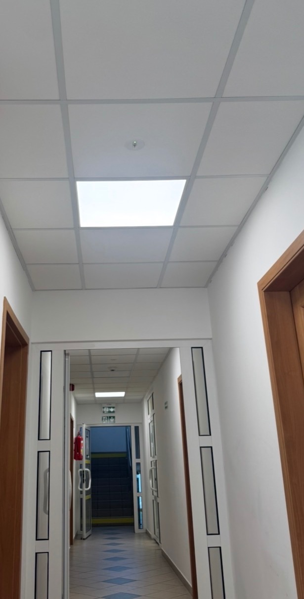 Korytarz biurowy z podwieszanym sufitem kasetonowym i wbudowanym oświetleniem LED. Widoczny czujnik dymu na suficie i drzwi po bokach korytarza.
