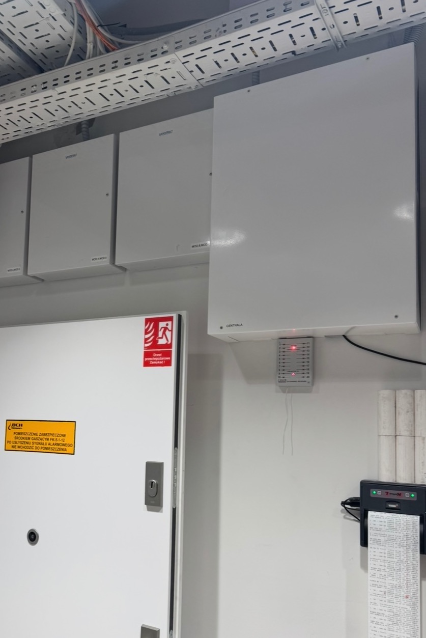 Białe szafy sterownicze alarmowe na ścianie, z widoczną centralą i drukarką raportów. Nad szafami koryta kablowe. Tabliczka ostrzegawcza na drzwiach.