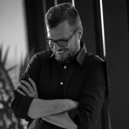 Czarno-białe zdjęcie uśmiechniętego mężczyzny w okularach, opierającego się o ścianę z założonymi rękami. Widoczna roślina w tle. Profesjonalny wizerunek.