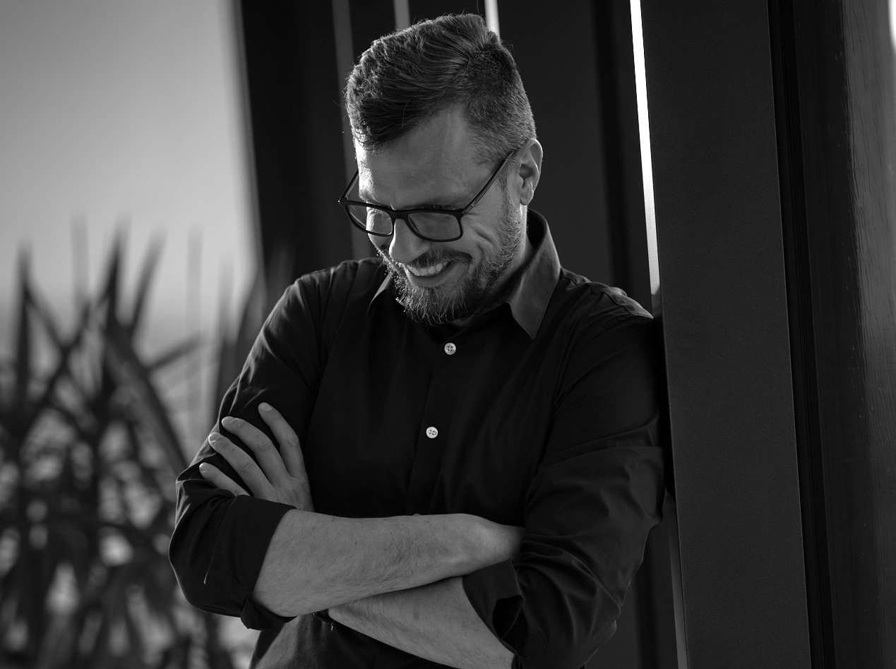 Czarno-białe zdjęcie uśmiechniętego mężczyzny w okularach, opierającego się o ścianę z założonymi rękami. Widoczna roślina w tle. Profesjonalny wizerunek.