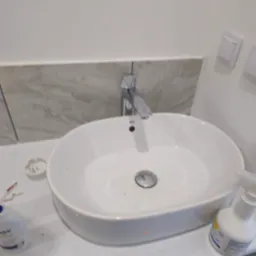 Biała umywalka z chromowaną baterią w trakcie montażu, widoczne narzędzia i akcesoria. Ujęcie z bliska, ukazujące detale instalacji w łazience.