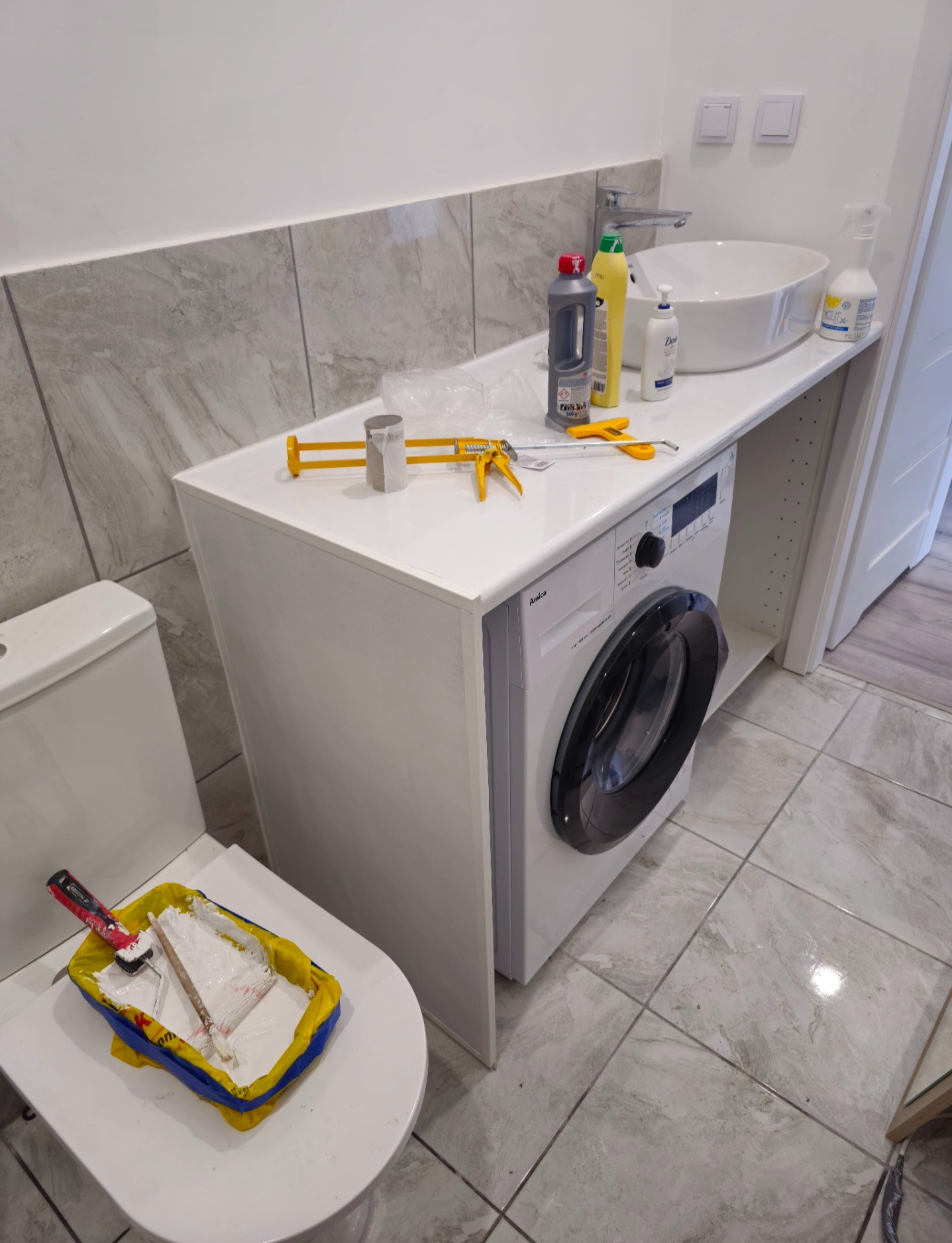 Biała łazienka z umywalką nablatową, pralką Amica i pojemnikiem z farbą na toalecie. Widoczne narzędzia i chemia gospodarcza. Płytki imitujące marmur.