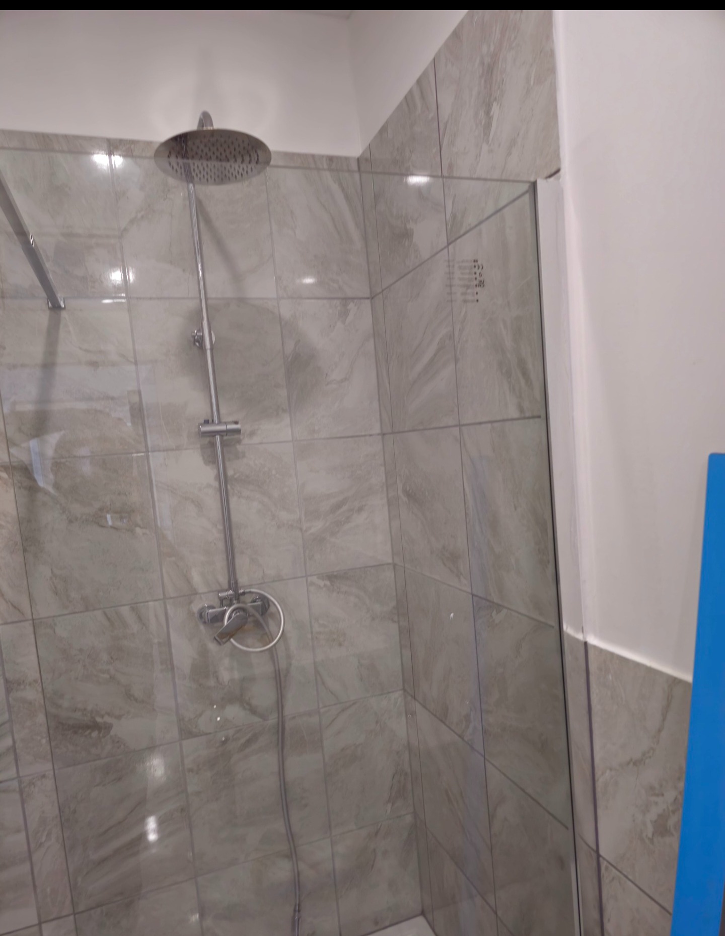 Wykończona kabina prysznicowa z dużym, okrągłym deszczownicą i szklaną ścianką. Płytki imitujące marmur na ścianach. Minimalistyczny design.