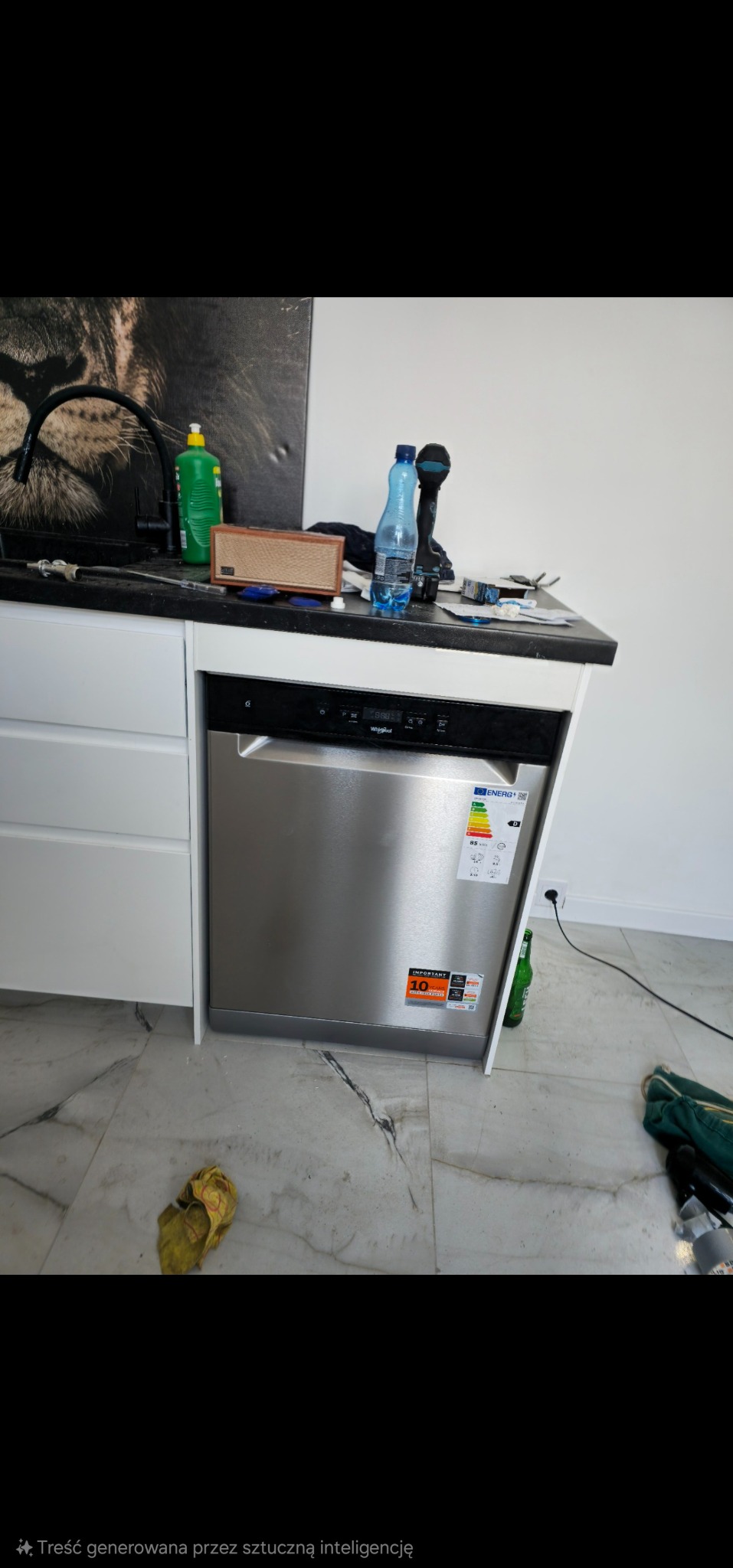 Nowoczesna kuchnia w trakcie montażu: srebrna zmywarka Whirlpool z naklejkami informacyjnymi, białe szafki, czarny blat, marmurowe płytki podłogowe i narzędzia.