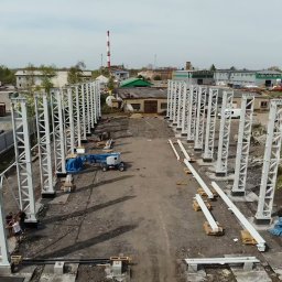 ZPUH INSTALATOR - Budowa hali: widok z góry na stalowe słupy konstrukcyjne, elementy na paletach i podnośnik koszowy. W tle budynki i komin.
