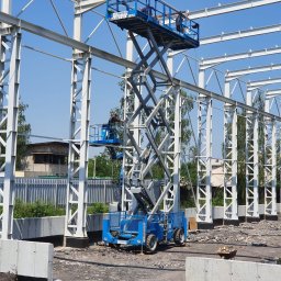 ZPUH INSTALATOR - Budowa hali w Wodzisławiu Śląskim: pracownicy na podnośniku montują stalową konstrukcję szkieletową na tle błękitnego nieba. Widoczne fundamenty.