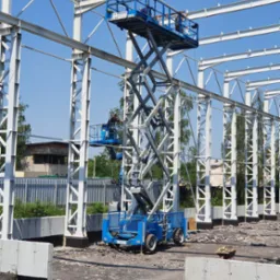 Budowa hali w Wodzisławiu Śląskim: pracownicy na podnośniku montują stalową konstrukcję szkieletową na tle błękitnego nieba. Widoczne fundamenty.