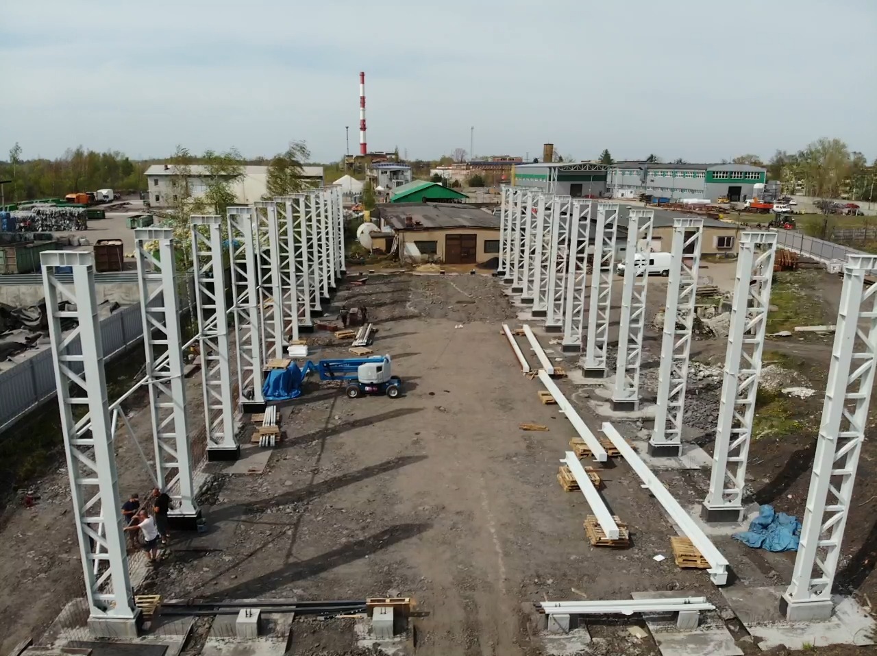 Budowa hali: widok z góry na stalowe słupy konstrukcyjne, elementy na paletach i podnośnik koszowy. W tle budynki i komin.