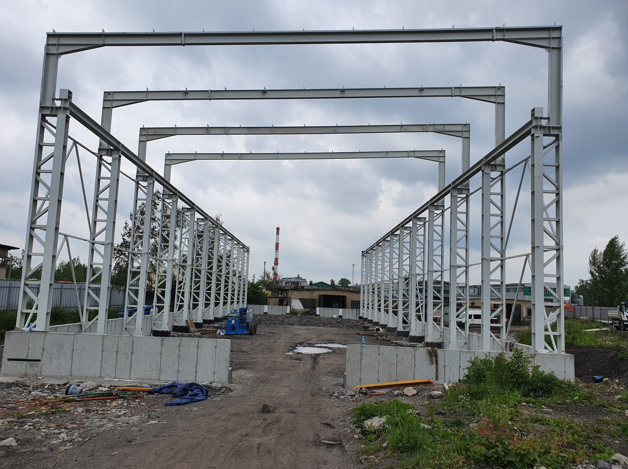 Wnętrze budowanej hali z widocznymi stalowymi ramami konstrukcyjnymi i betonowymi fundamentami, na tle pochmurnego nieba. Widoczne elementy placu budowy.