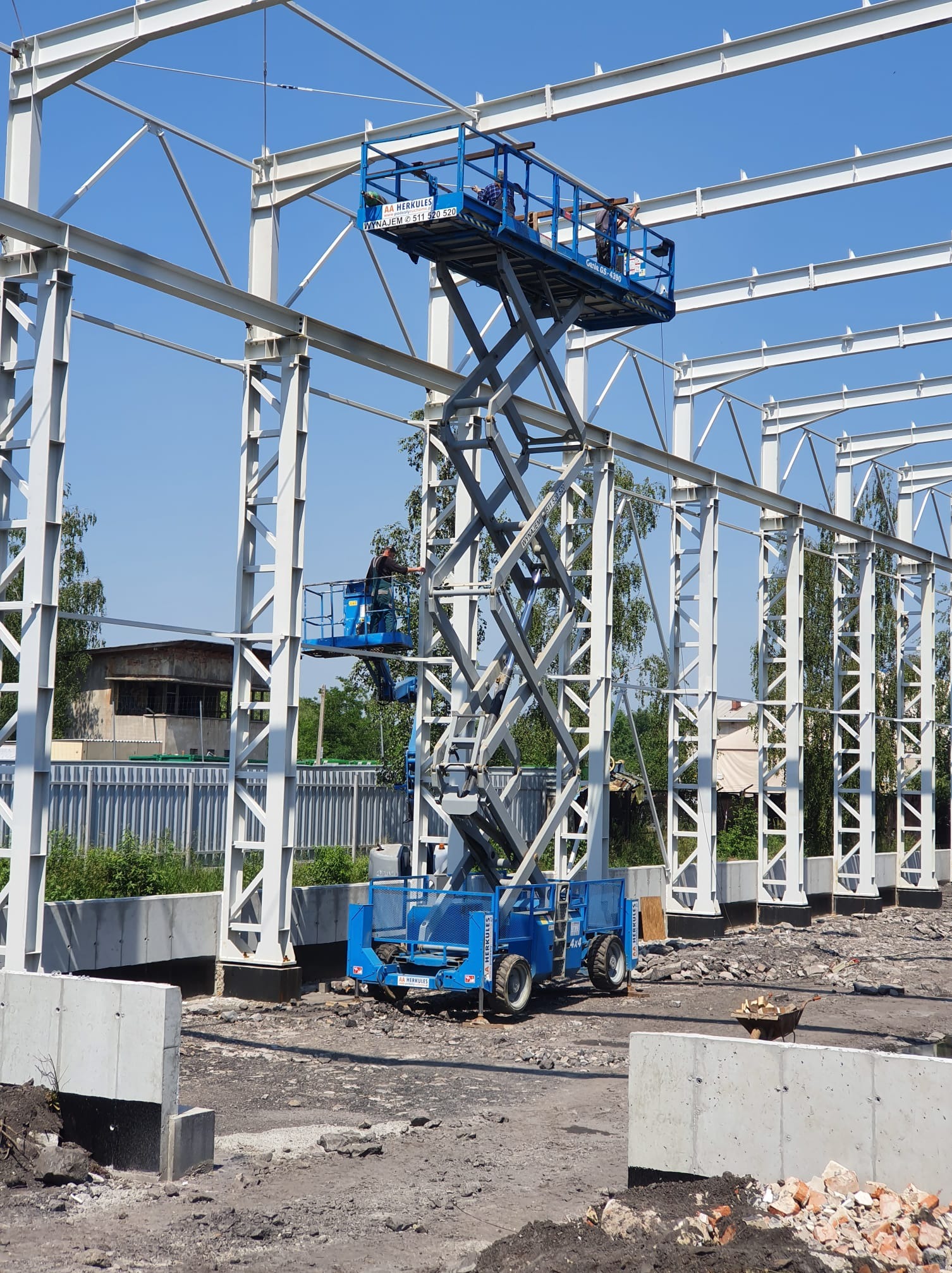 Budowa hali w Wodzisławiu Śląskim: pracownicy na podnośniku montują stalową konstrukcję szkieletową na tle błękitnego nieba. Widoczne fundamenty.