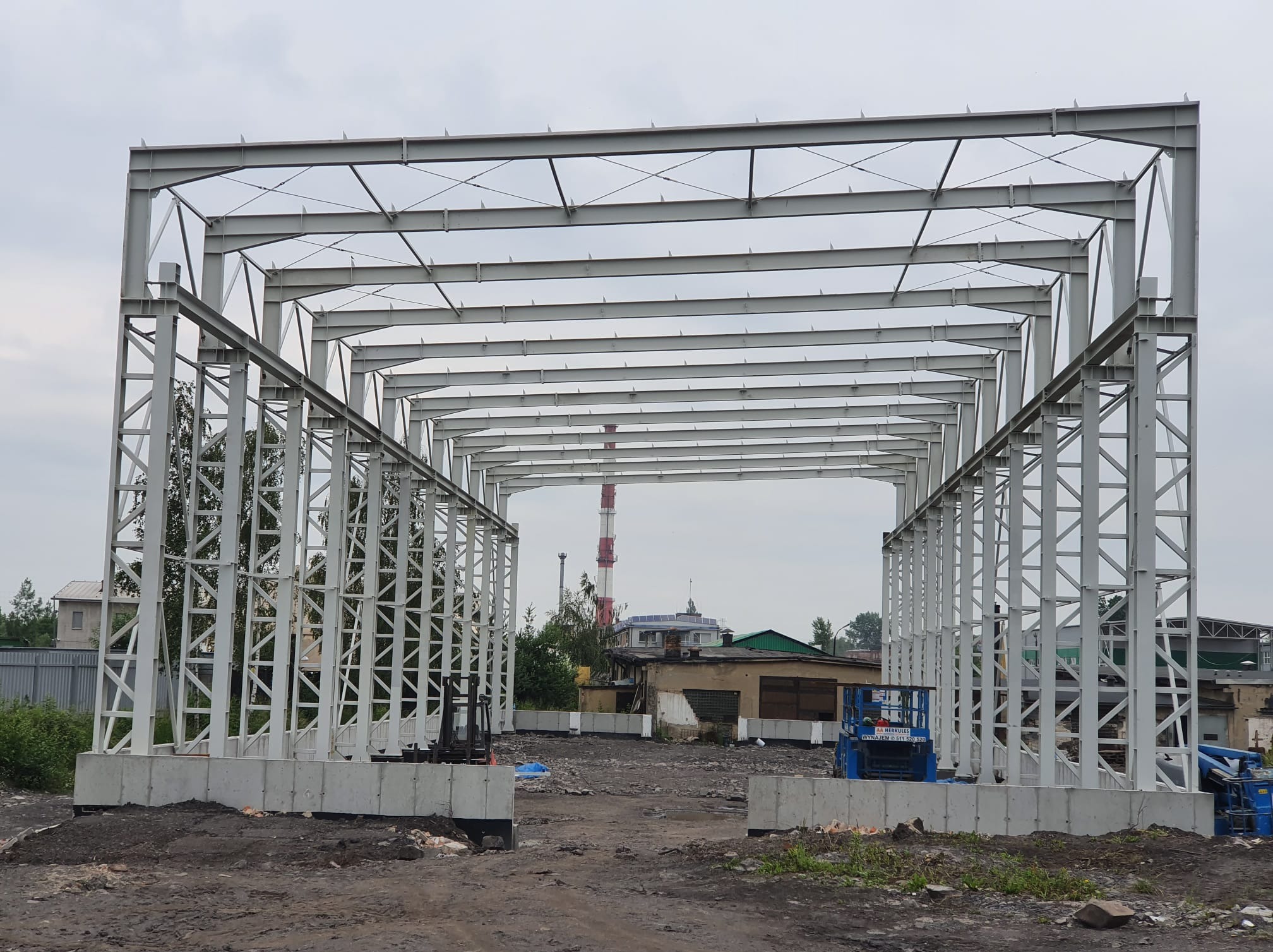 Konstrukcja stalowa hali w trakcie budowy, widoczne szkieletowe elementy, w tle komin i budynki. Ujęcie z poziomu gruntu, pochmurne niebo.