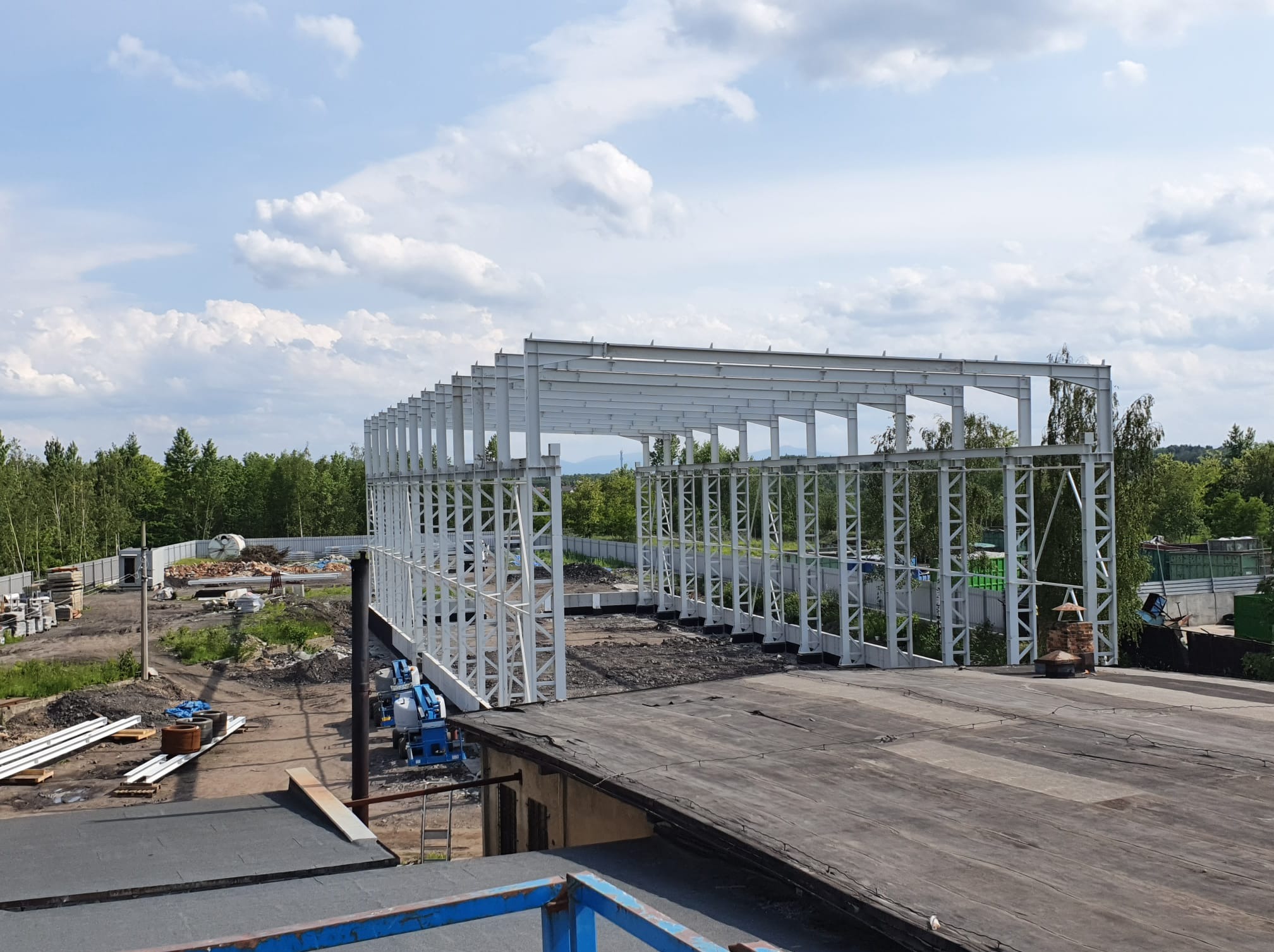 Konstrukcja stalowa hali w trakcie budowy, widok z góry na szkielet budynku z białych profili, z zielenią w tle i niebieskim niebem z chmurami.