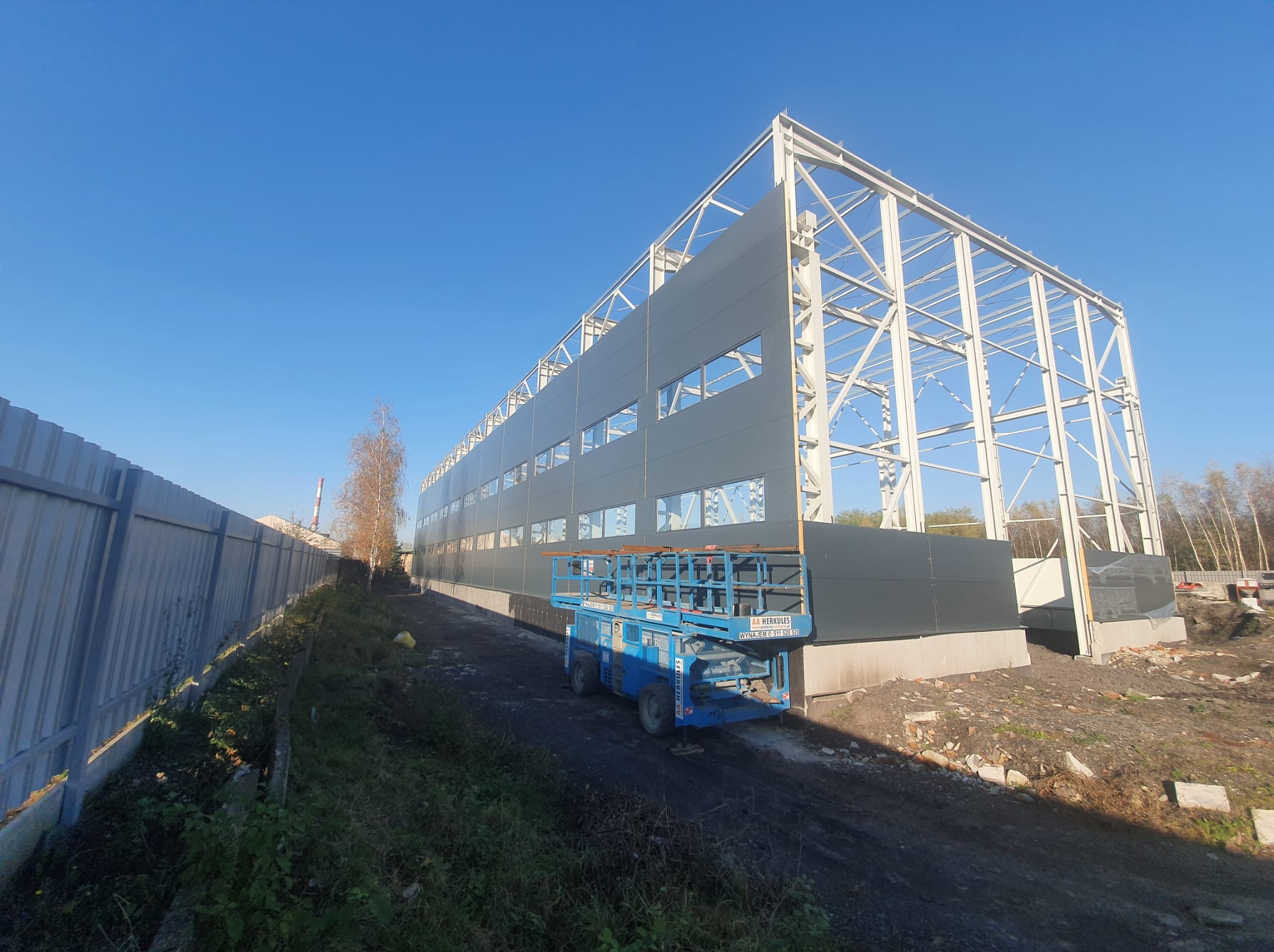 Montaż szarej płyty warstwowej na stalowej konstrukcji hali, widoczny podnośnik koszowy na tle błękitnego nieba. Częściowo ukończona elewacja.