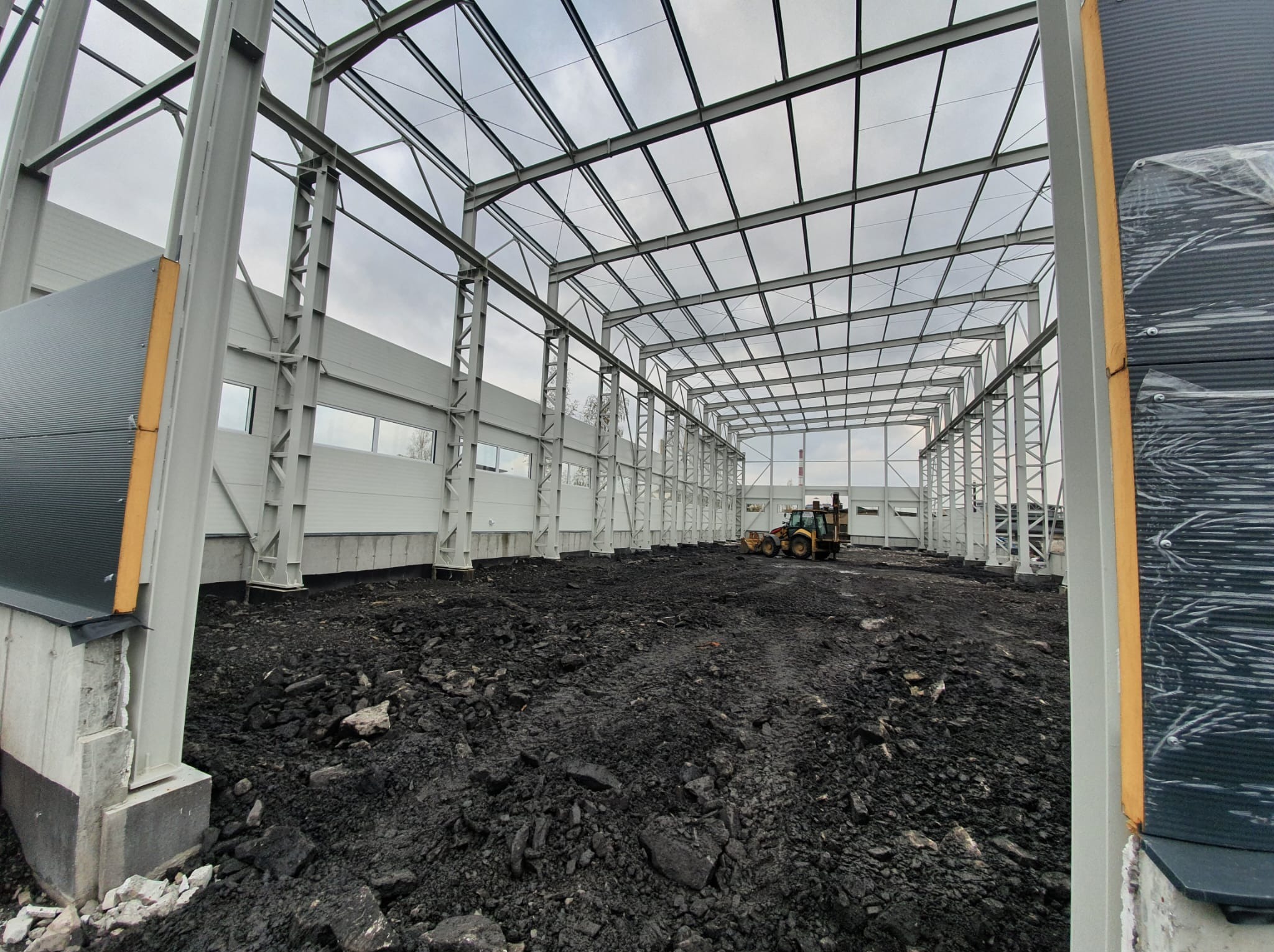 Budowa hali: stalowa konstrukcja szkieletowa z widoczną blachą trapezową i traktorem w tle, prezentująca postęp prac na placu budowy.