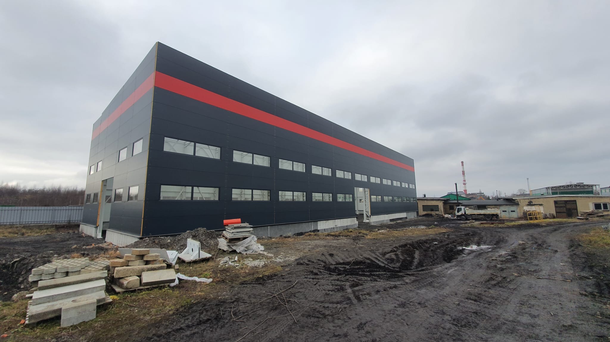 Nowoczesna hala przemysłowa z czarną elewacją i czerwonym pasem, widoczne okna i fragment placu budowy. Pochmurne niebo w tle.