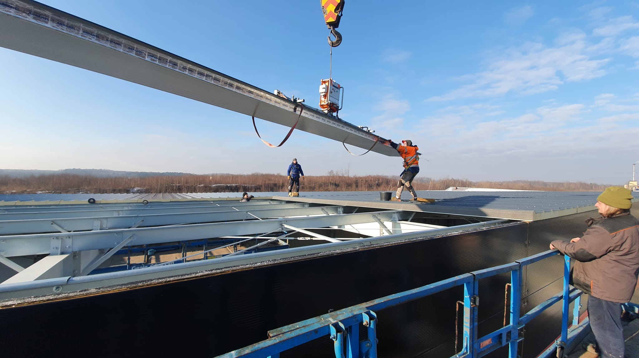 Montaż płyty warstwowej na konstrukcji hali. Pracownicy w kaskach i uprzężach, dźwig w tle, widok na okolicę. Praca na wysokościach, bezpieczeństwo.
