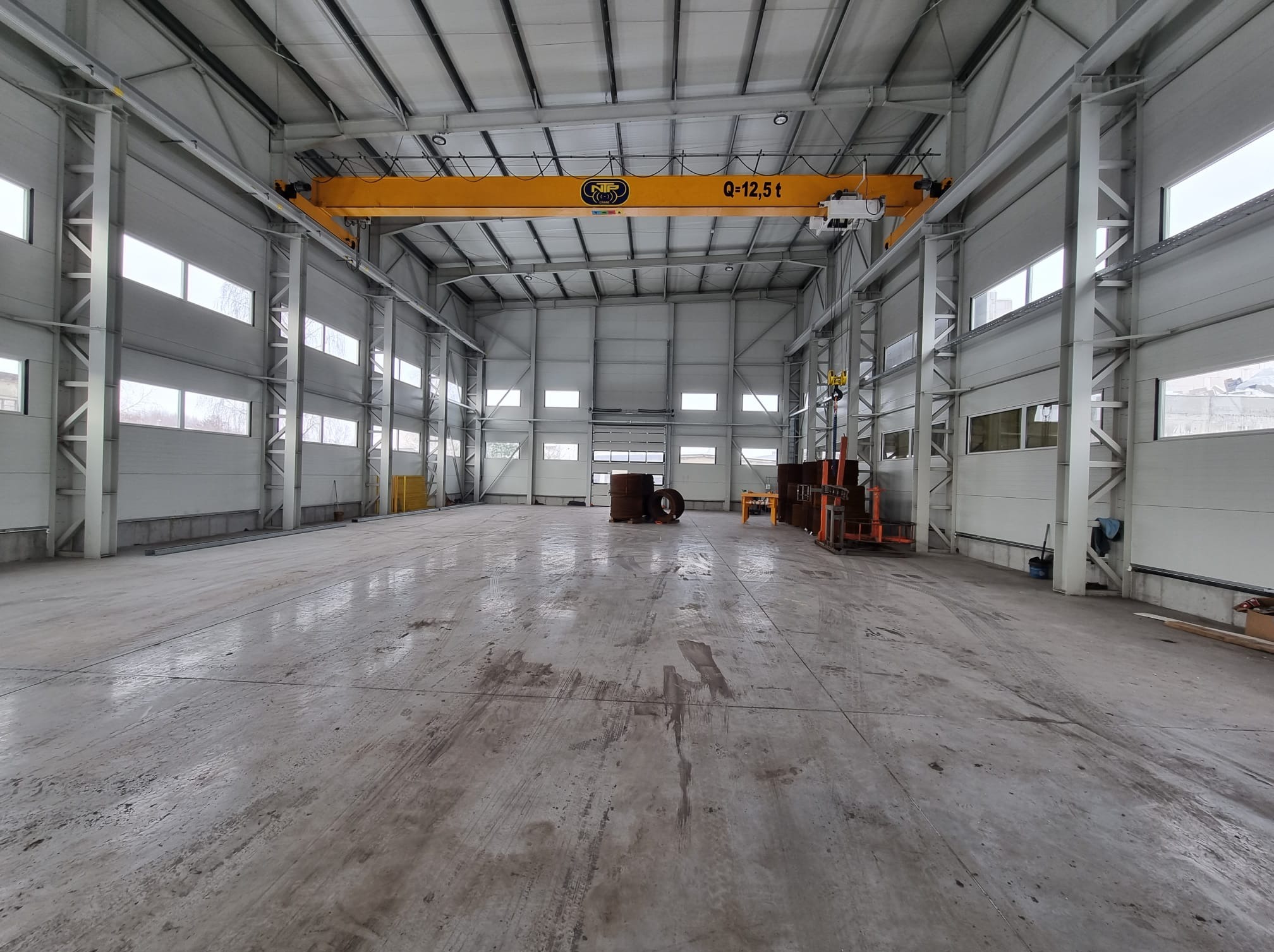 Wnętrze hali produkcyjnej z suwnicą. Widoczne stalowe konstrukcje, okna i brama wjazdowa. Podłoga betonowa z widocznymi śladami użytkowania.