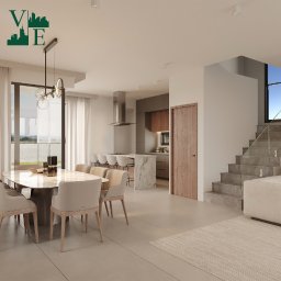 Velora Estate - Biuro Nieruchomości - Jasne wnętrze domu z marmurowym stołem jadalnym, kuchnią z wyspą i schodami. Nowoczesny design z widokiem na zewnątrz przez duże okno.