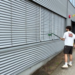 Mycie okien - Poznań - Pracownik czyści żaluzje na elewacji budynku przemysłowego za pomocą szczotki na długim kiju. Obok widoczna betonowa podmurówka i chodnik z kostki.