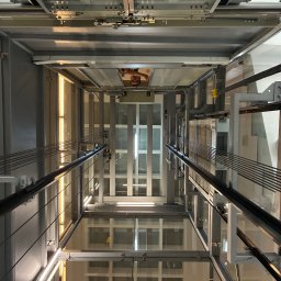 Mycie okien - Poznań - Wnętrze szybu windy z perspektywy pionowej, ukazujące konstrukcję metalową, szklane panele i oświetlenie. Widoczny fragment osoby w górnej części ujęcia.