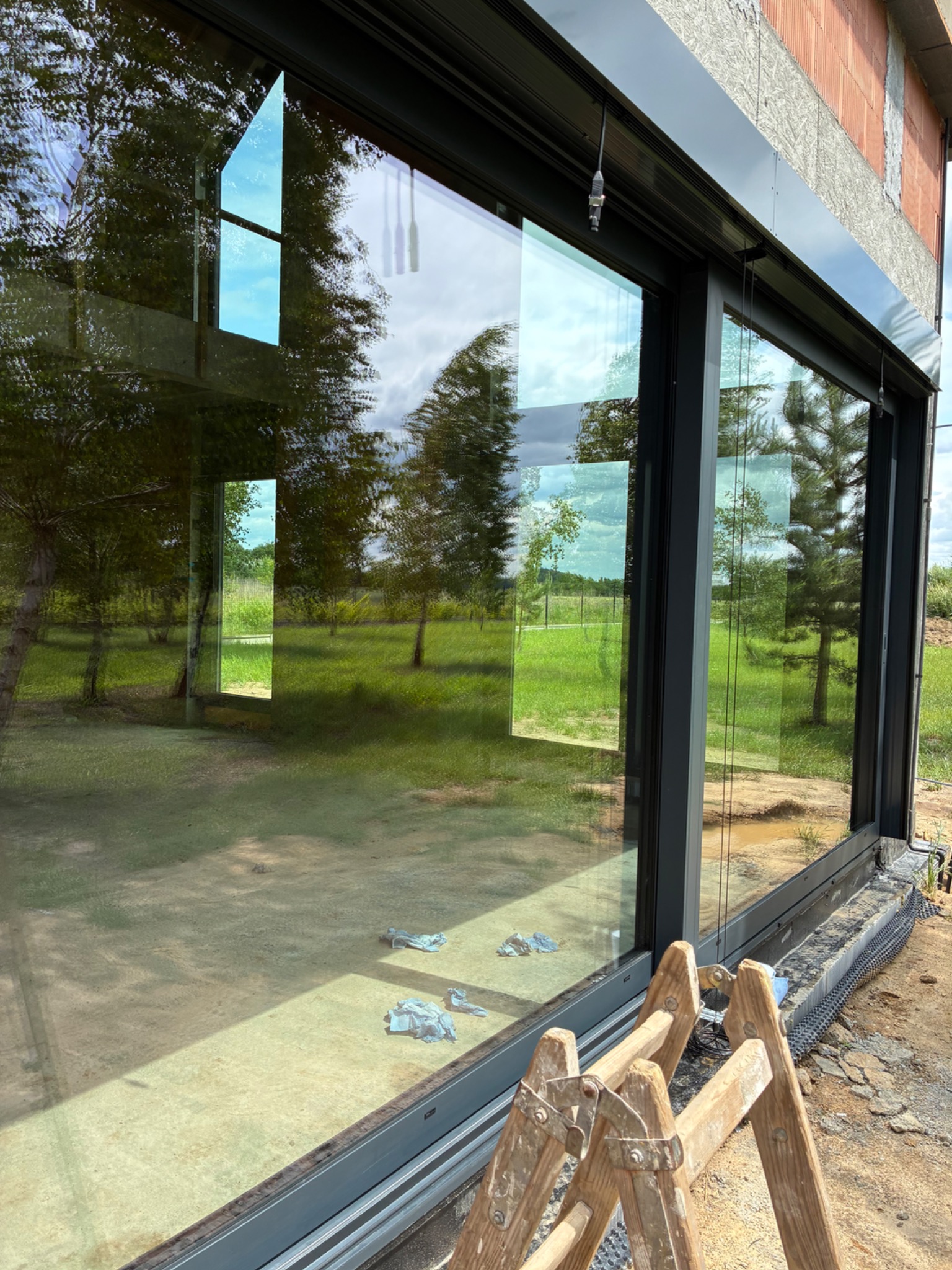 Duże okno z czarną ramą, odbijające drzewa i niebo. Na dole drabina i ślady po czyszczeniu. Widoczny fragment elewacji w stanie surowym.