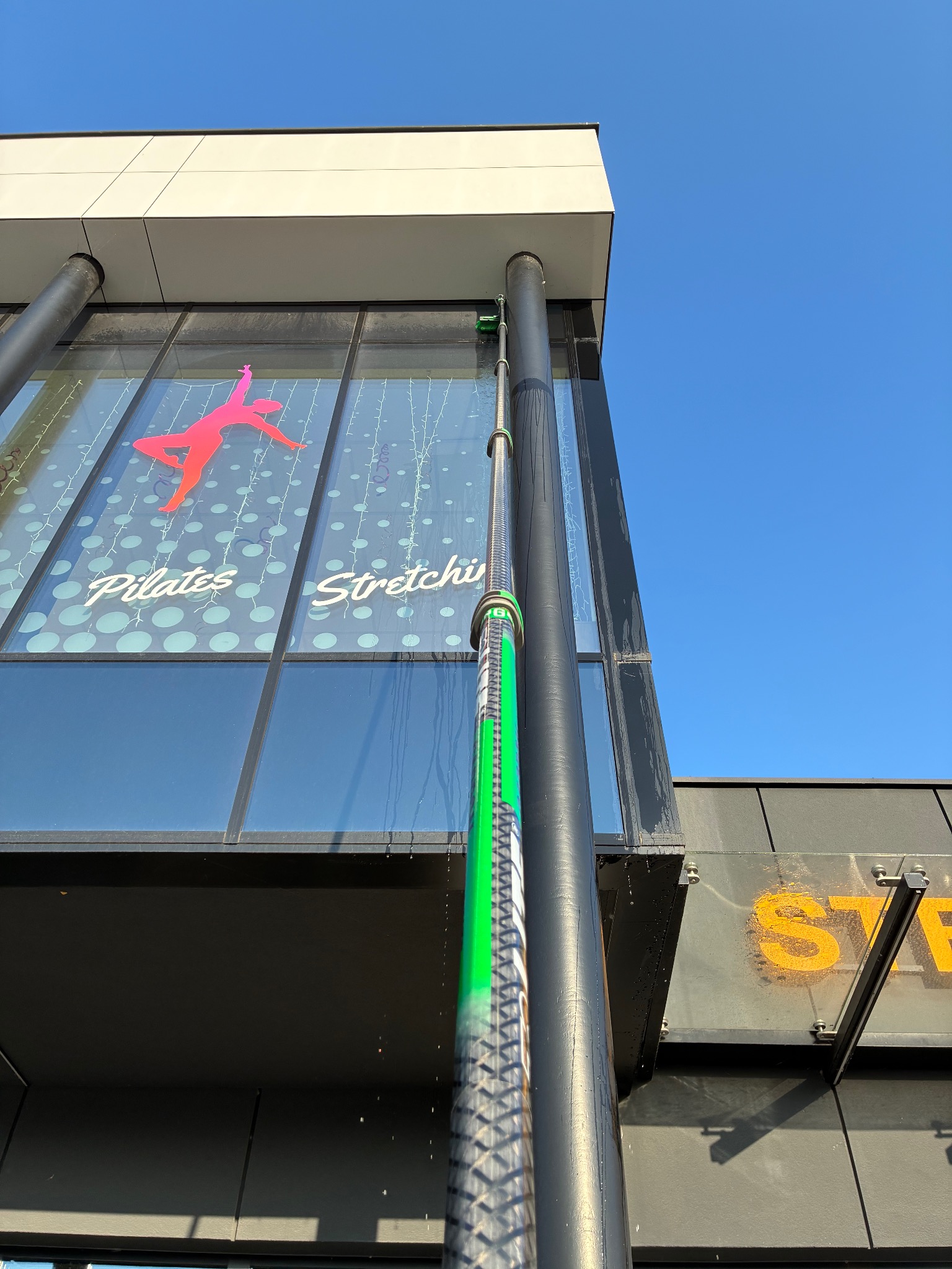 Mycie okien na wysokości budynku z reklamą Pilates Stretch za pomocą specjalistycznego sprzętu teleskopowego. Widoczne ściekające krople wody.