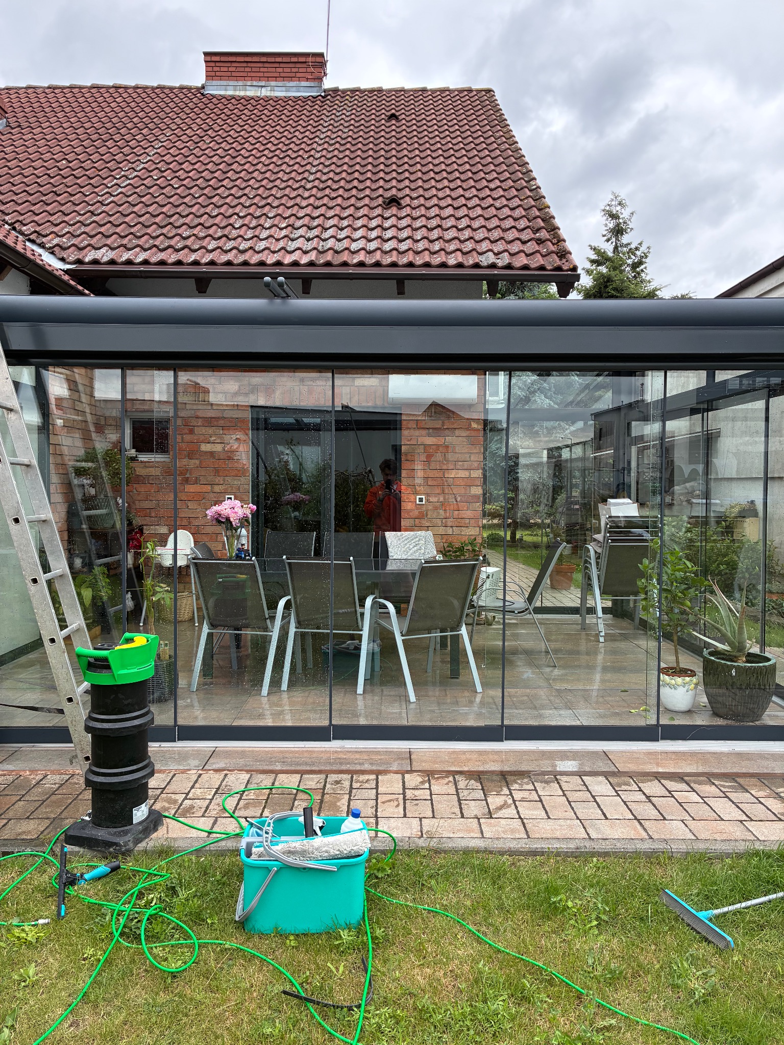 Szklana weranda po umyciu, widoczne narzędzia i drabina. Wnętrze z kwiatami i meblami ogrodowymi. Czyste szyby odbijają otoczenie.