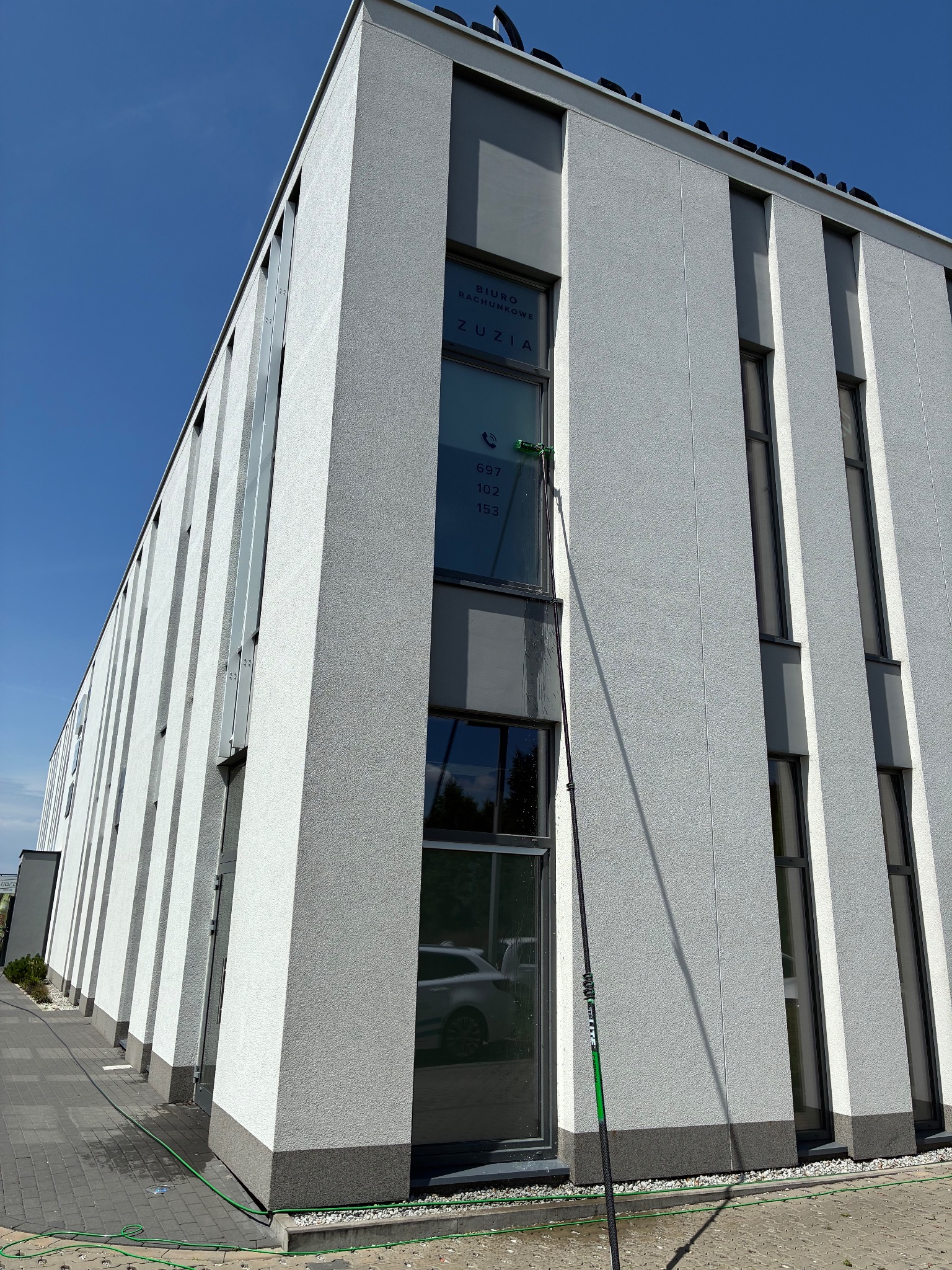 Mycie okien biurowca z użyciem lancy teleskopowej. Na oknie widoczna reklama biura rachunkowego. W tle błękitne niebo i fragment chodnika z wężem ogrodowym.