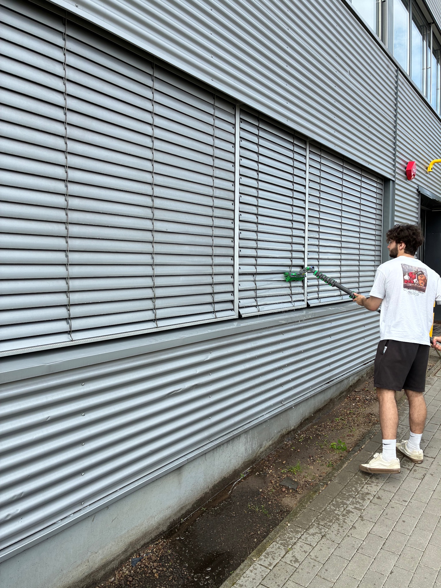 Pracownik czyści żaluzje na elewacji budynku przemysłowego za pomocą szczotki na długim kiju. Obok widoczna betonowa podmurówka i chodnik z kostki.