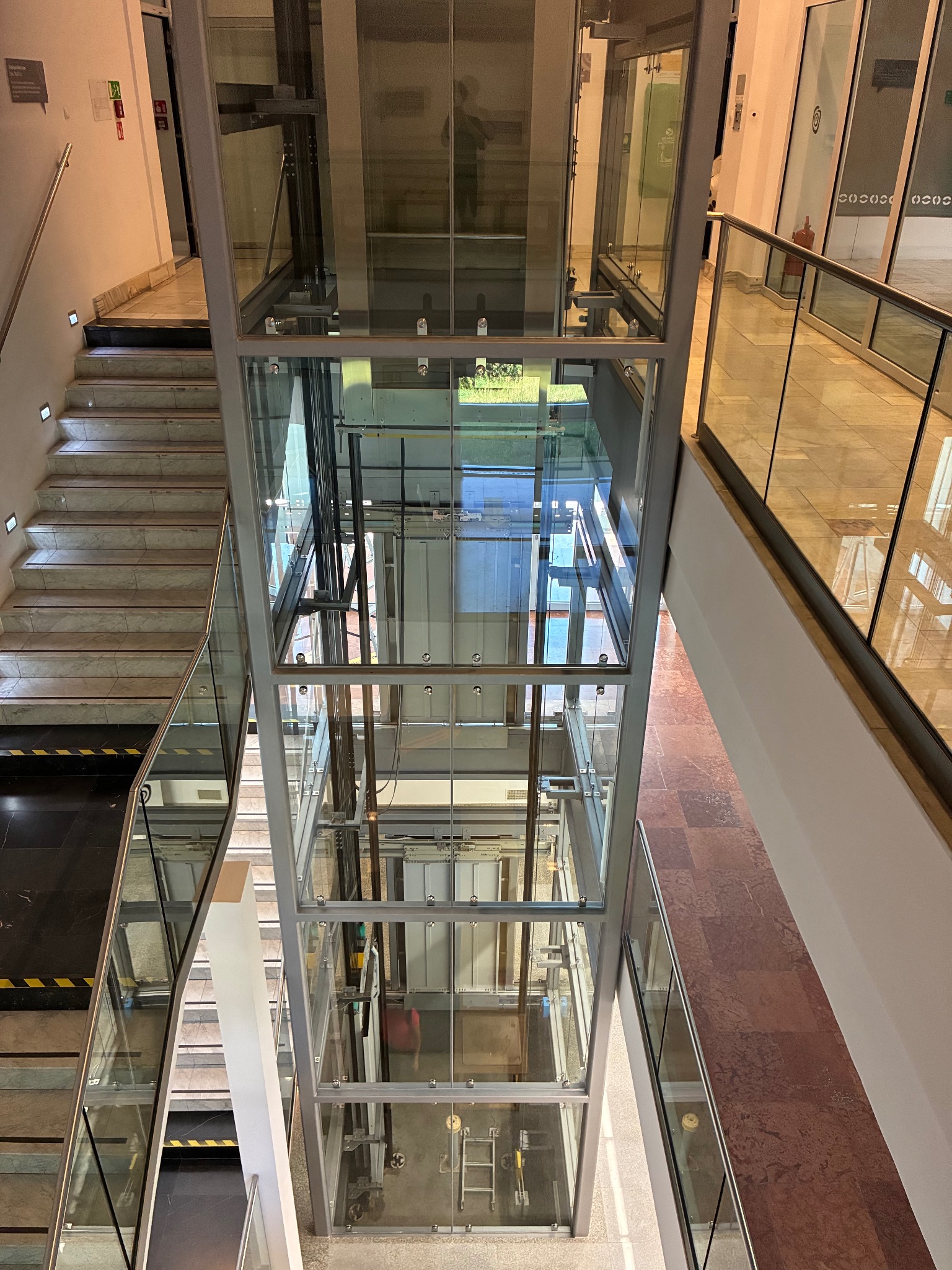 Szklana winda w budynku biurowym, widok z góry. Widoczne schody, balustrady i fragmenty korytarzy. Przejrzysta konstrukcja windy pozwala obserwować jej mechanizm.