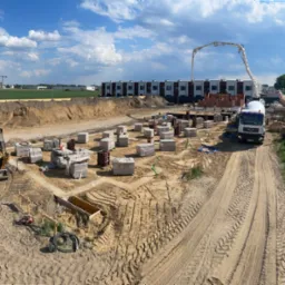 Szeroki kadr na plac budowy z fundamentami pod budynek, widoczny betonowóz z pompą, koparka oraz ułożone bloczki fundamentowe. Słoneczny dzień.