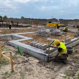 RENOMA INWESTYCJE SPÓŁKA Z OGRANICZONĄ ODPOWIEDZIALNOŚCIĄ - Budowa fundamentów domu: pracownicy układają bloczki betonowe na przygotowanym podłożu, używając poziomicy. W tle widoczne materiały budowlane i wykop pod fundamenty.