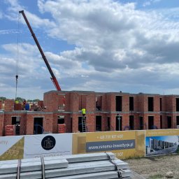 RENOMA INWESTYCJE SPÓŁKA Z OGRANICZONĄ ODPOWIEDZIALNOŚCIĄ - Budowa domów z czerwonej cegły, widoczny dźwig i pracownicy w żółtych kamizelkach. Na pierwszym planie baner reklamowy z wizualizacją osiedla.