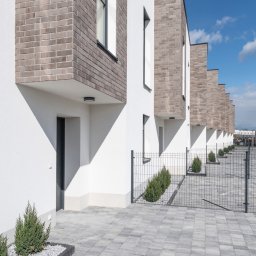 RENOMA INWESTYCJE SPÓŁKA Z OGRANICZONĄ ODPOWIEDZIALNOŚCIĄ - Szereg identycznych, nowoczesnych domów z białymi ścianami i ceglanymi elementami, widok z zewnątrz, z kostką brukową i niskimi krzewami wzdłuż ogrodzenia.