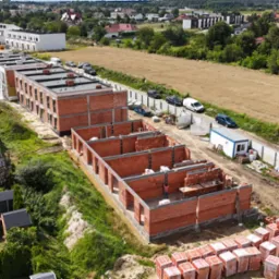 Budowa osiedla domów z czerwonej cegły, widok z góry. Etap murowania ścian, widoczne palety z cegłami i pracownik wewnątrz konstrukcji.