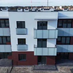 Nowoczesny, trzypiętrowy budynek z balkonami o szklanych balustradach, wykończony cegłą i białym tynkiem. Widok z góry, w tle zabudowa miejska i pole.