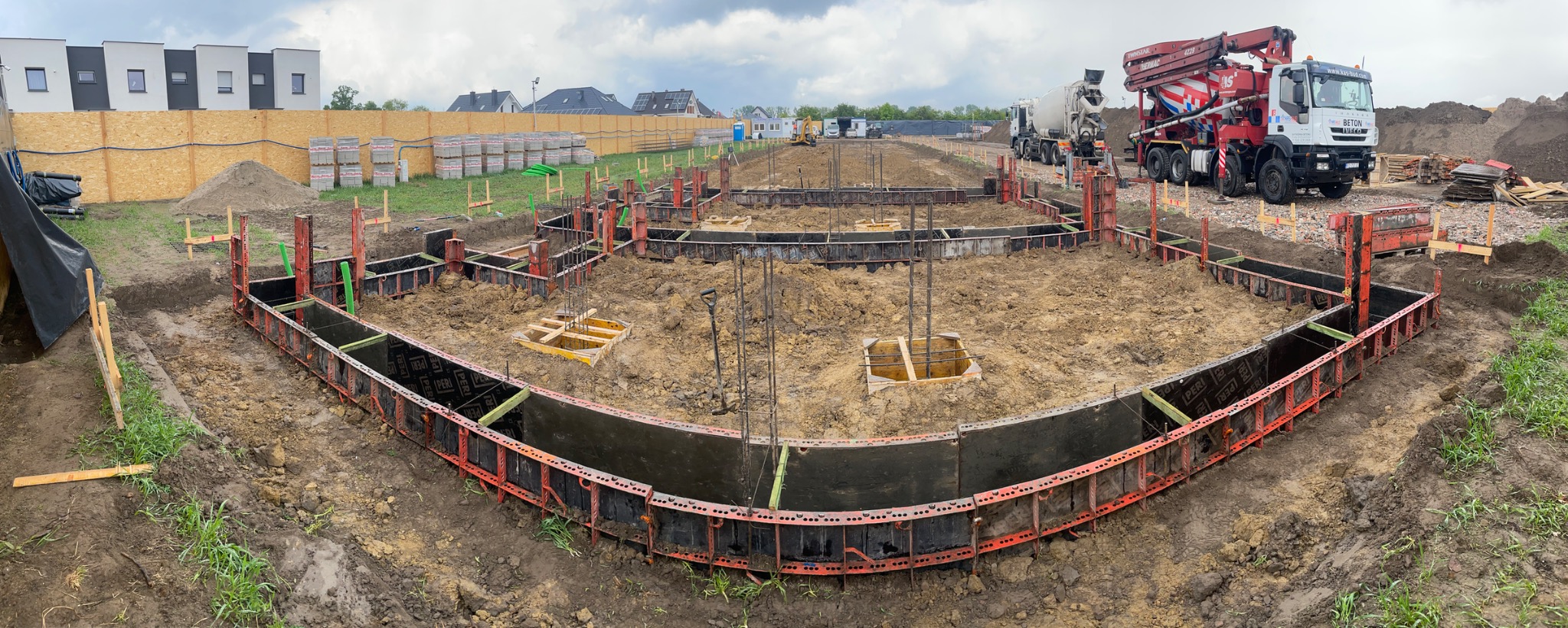 Szeroki kadr na wykop z szalunkami fundamentowymi, zbrojeniami i betonowozem w tle, na placu budowy w trakcie prac przygotowawczych.