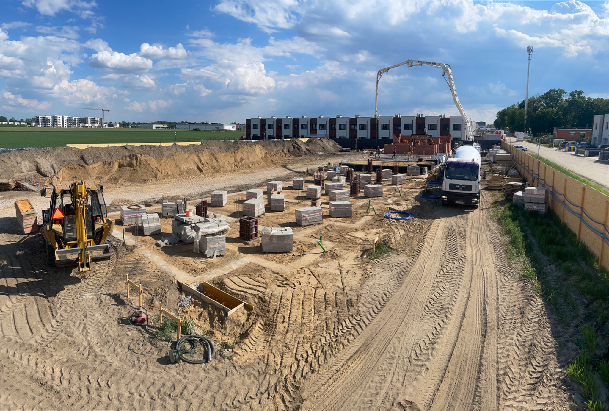 Szeroki kadr na plac budowy z fundamentami pod budynek, widoczny betonowóz z pompą, koparka oraz ułożone bloczki fundamentowe. Słoneczny dzień.