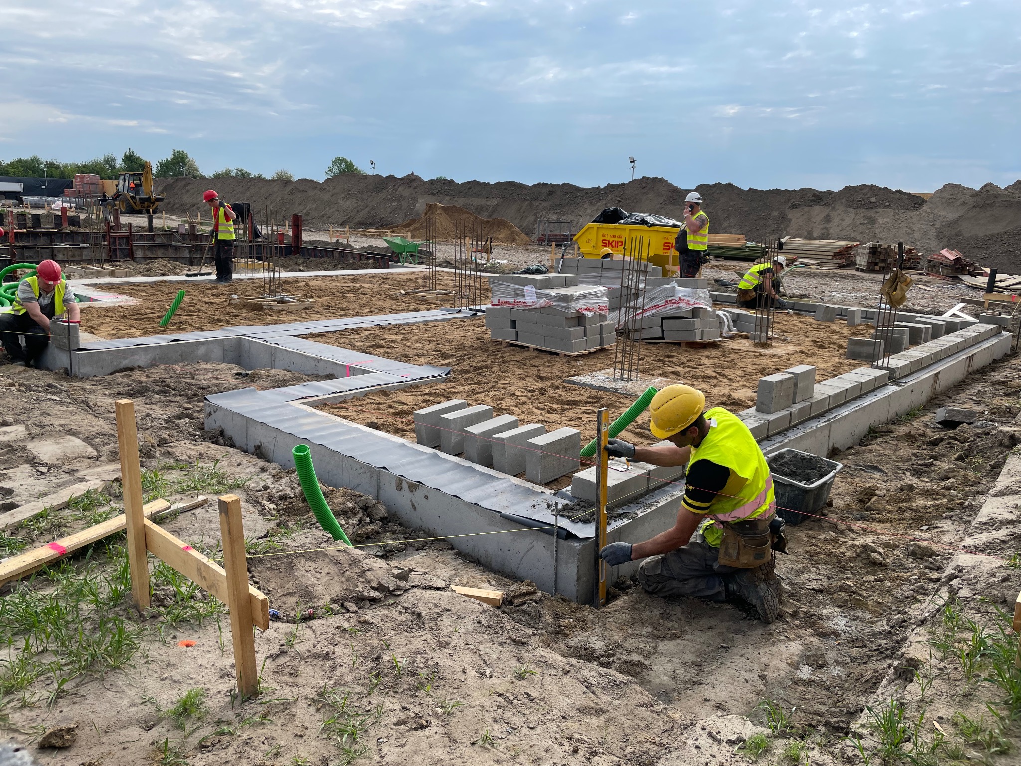 Budowa fundamentów domu: pracownicy układają bloczki betonowe na przygotowanym podłożu, używając poziomicy. W tle widoczne materiały budowlane i wykop pod fundamenty.