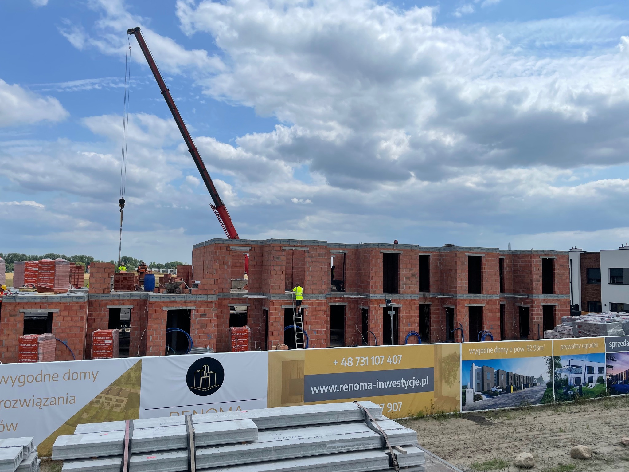 Budowa domów z czerwonej cegły, widoczny dźwig i pracownicy w żółtych kamizelkach. Na pierwszym planie baner reklamowy z wizualizacją osiedla.