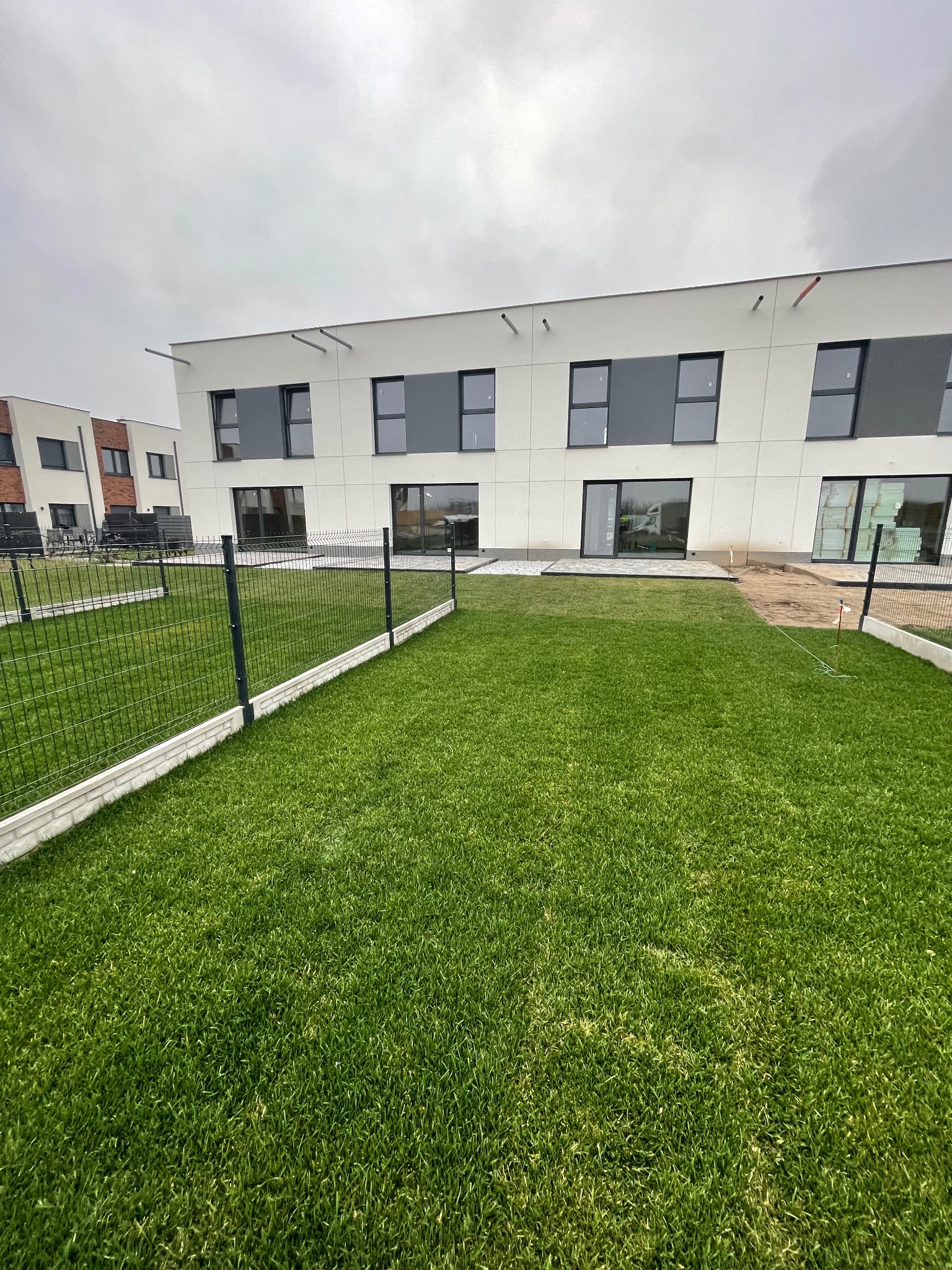 Nowoczesny dom z ogrodem i metalowym ogrodzeniem panelowym. Widoczne okna, drzwi balkonowe i trawnik. Pochmurne niebo.