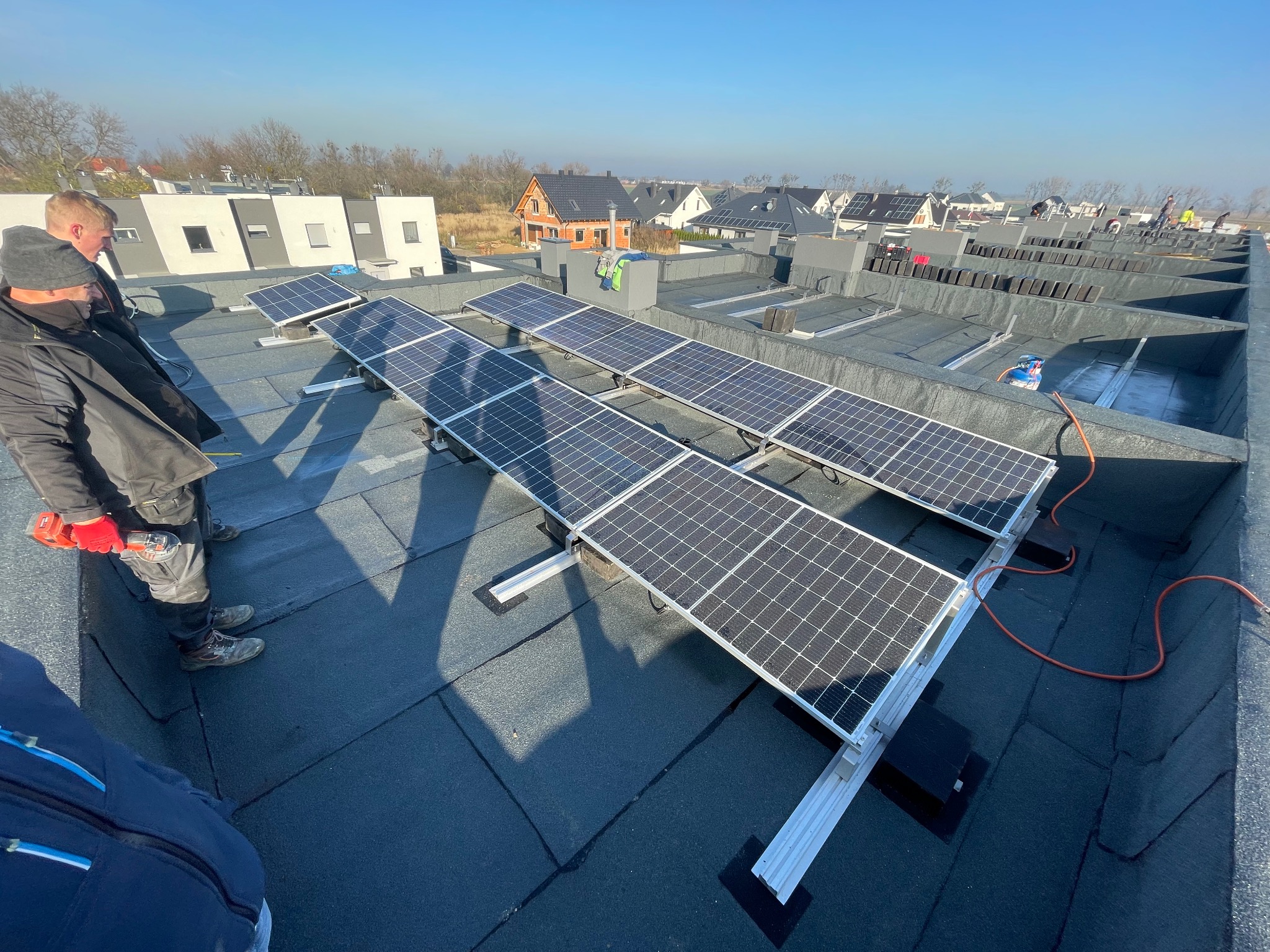 Montaż paneli słonecznych na płaskim dachu budynku. Widoczni dwaj monterzy podczas pracy. W tle zabudowa jednorodzinna i błękitne niebo.
