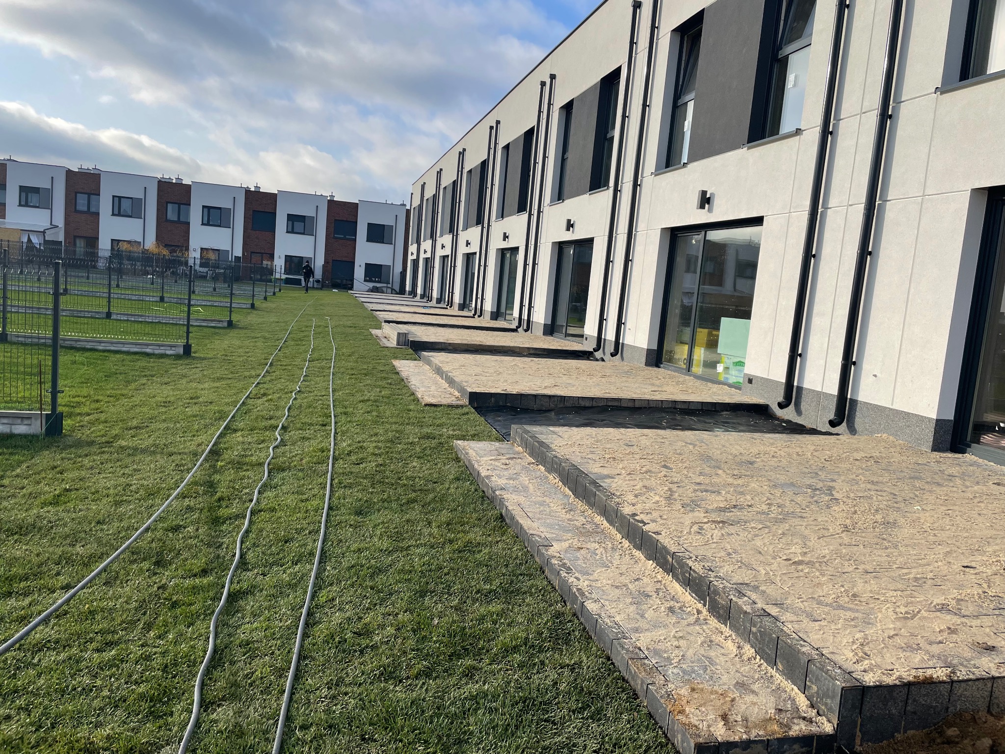 Nowoczesny szereg domów z wybrukowanymi tarasami i trawnikiem. Widoczne rury na trawie, prawdopodobnie instalacja nawadniająca. Ujęcie z perspektywy trawnika.