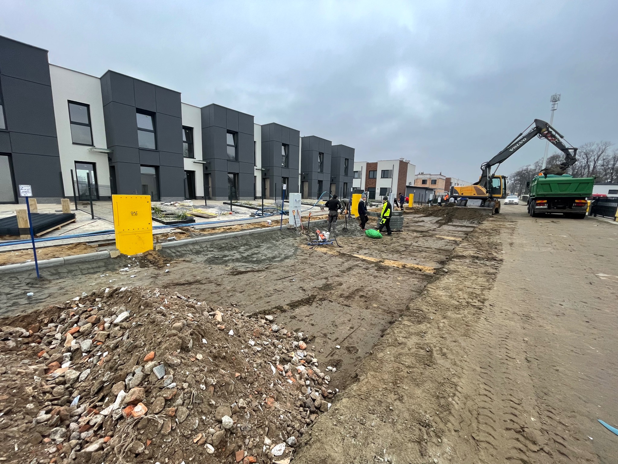 Prace ziemne na budowie osiedla: robotnicy, koparka ładująca gruz do wywrotki, w tle nowoczesne domy szeregowe z szarymi elewacjami i żółta skrzynka gazowa.