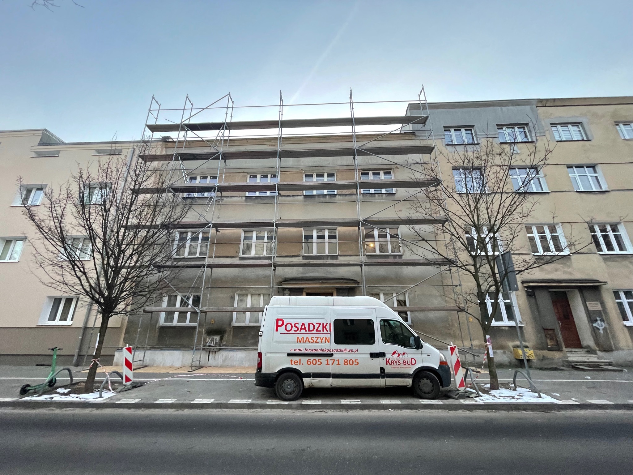Budynek w trakcie remontu elewacji, otoczony rusztowaniami. Na pierwszym planie zaparkowany biały bus z reklamą firmy budowlanej i posadzek.